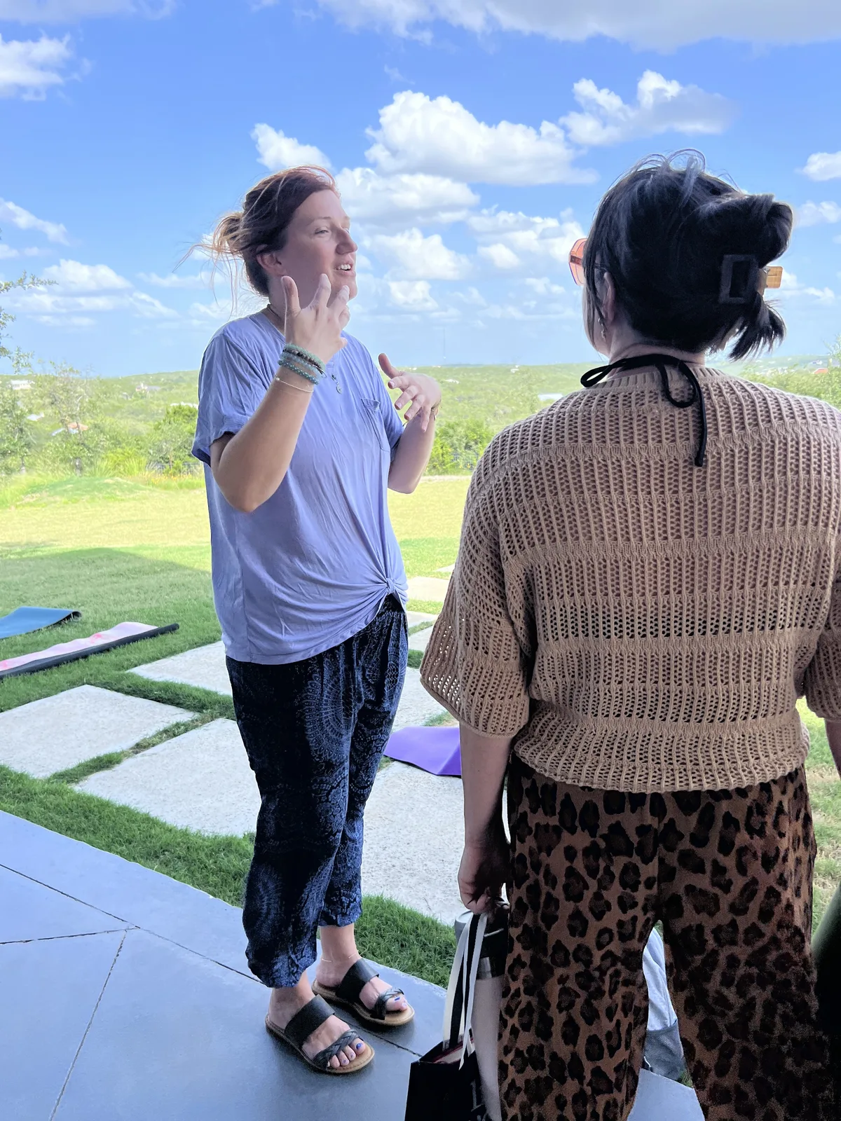 Two friends laughing and talking outdoors near their yoga mats under a blue sky