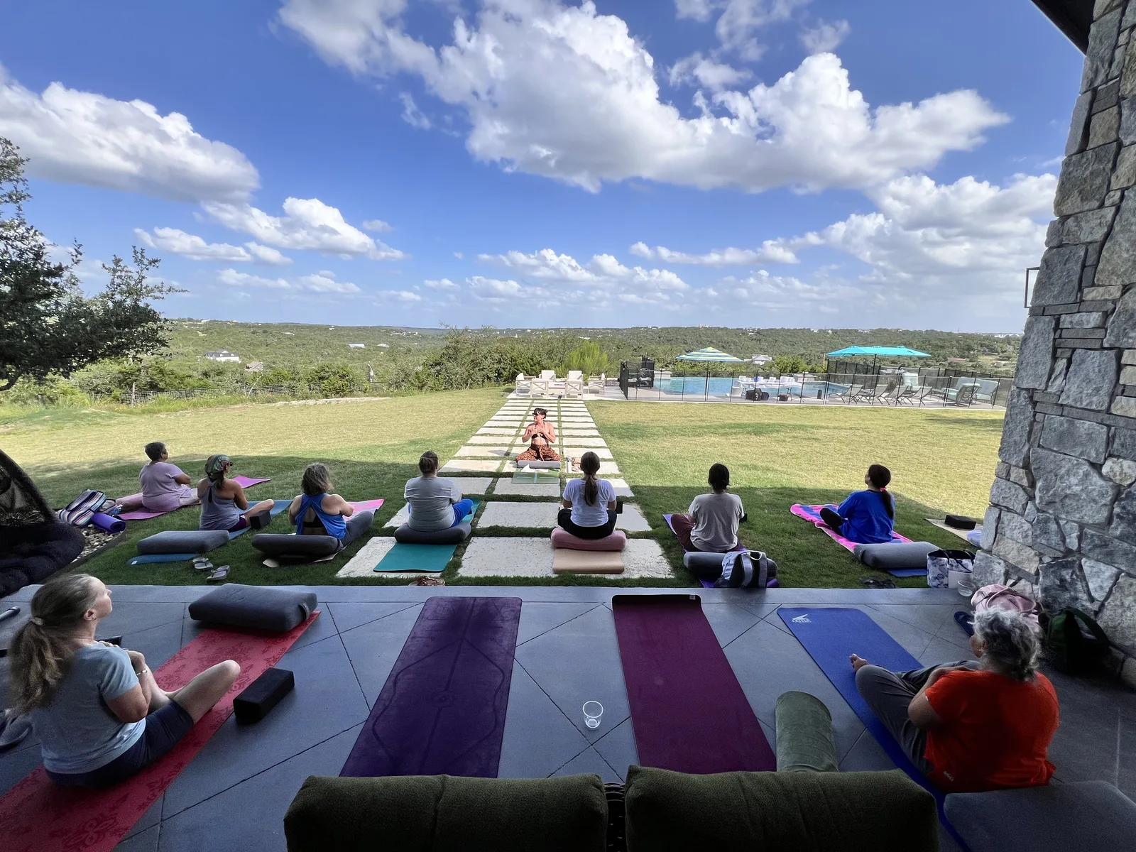 A full outdoor yoga class seated on mats overlooking the Hill Country, with the pool and patio behind them