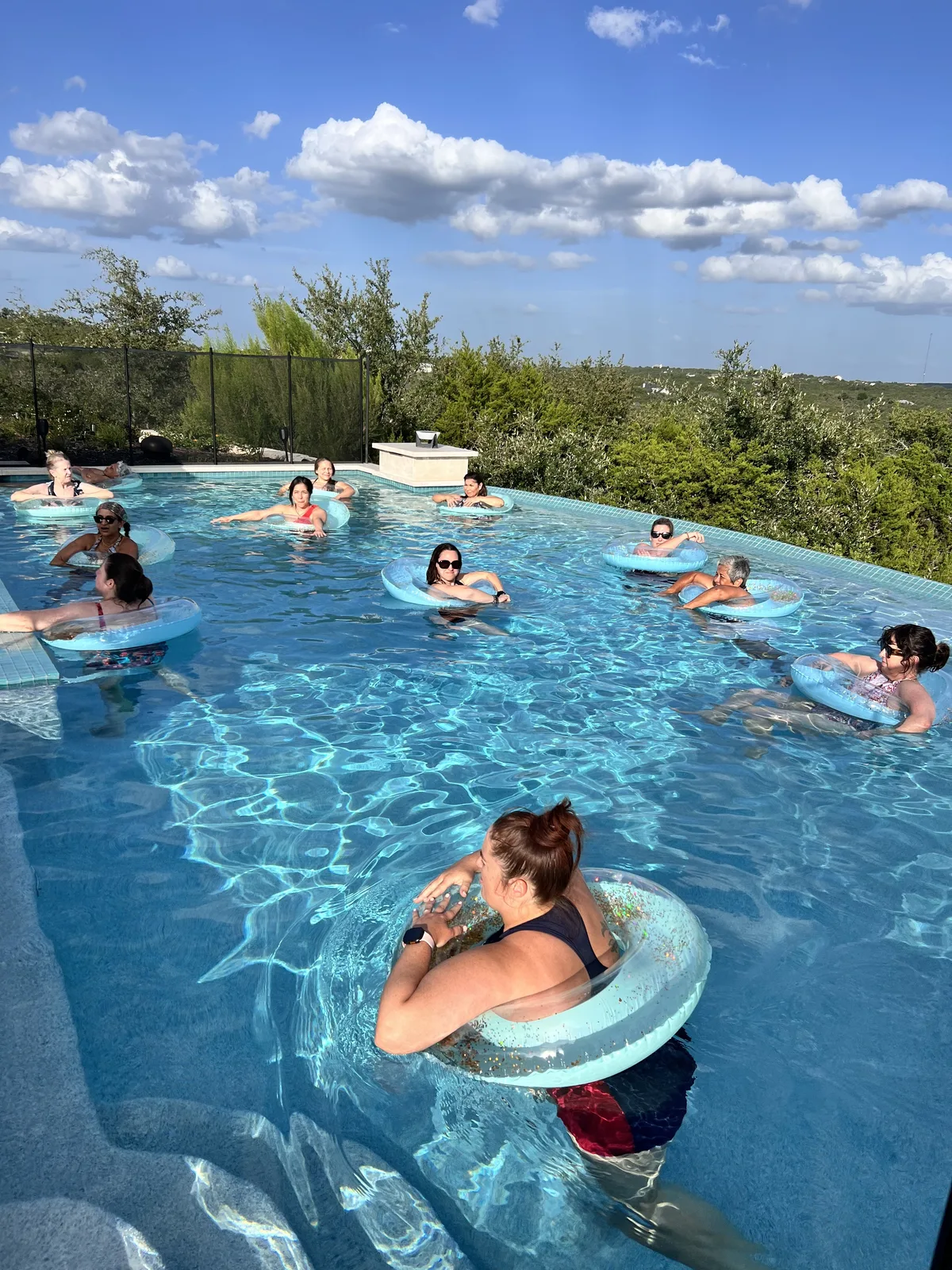 A group floating in rings in an infinity pool during a poolside sound bath, Hill Country hills in the distance